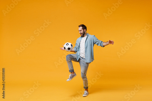 Cheerful young man in casual blue shirt posing isolated on yellow orange wall background  studio portrait. People emotions lifestyle concept. Mock up copy space. Juggling bouncing soccer ball on knee.