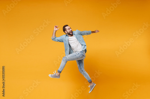 Side view of cheerful young bearded man in casual blue shirt posing isolated on yellow orange background  studio portrait. People emotions lifestyle concept. Mock up copy space. Jumping looking back.