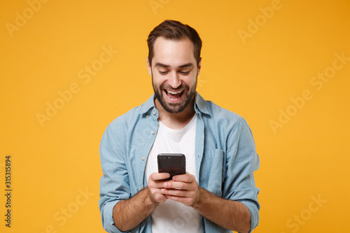 Cheerful young man in casual blue shirt posing isolated on yellow orange wall background studio portrait. People emotions lifestyle concept. Mock up copy space. Using mobile phone, typing sms message.
