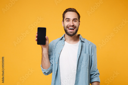 Laughing young man in casual blue shirt posing isolated on yellow orange wall background, studio portrait. People lifestyle concept. Mock up copy space. Holding mobile phone with blank empty screen.