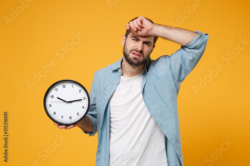 Tired young man in casual blue shirt posing isolated on yellow orange background, studio portrait. People sincere emotions lifestyle concept. Mock up copy space. Hold in hands clock, put hand on head.