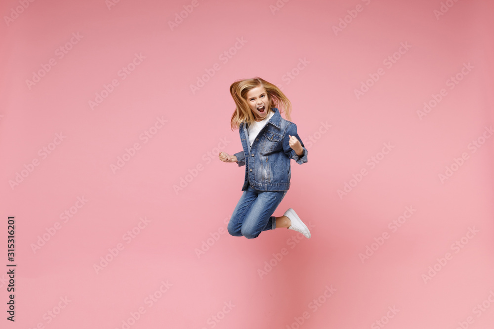 Screaming little blonde kid girl 12-13 years old in denim jacket isolated on pastel pink background children portrait. Childhood lifestyle concept. Mock up copy space. Jumping, doing winner gesture.