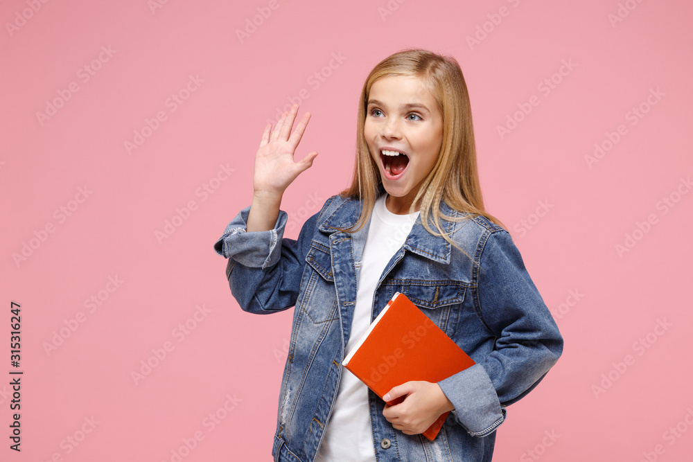 Excited little kid girl 12-13 years old in denim jacket isolated on pastel  pink background. Childhood lifestyle concept. Mock up copy space. Hold  notebook waving greeting with hand as notices someone. foto