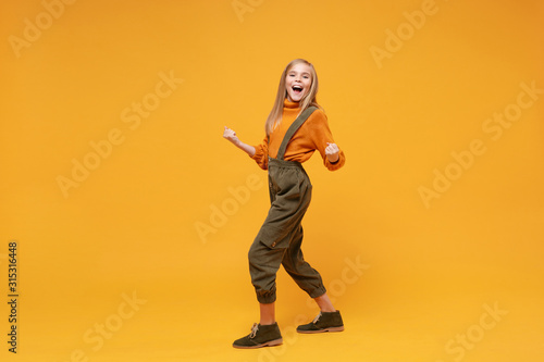 Happy little blonde kid girl 12-13 years old in turtleneck, jumpsuit isolated on orange yellow wall background in studio. Childhood lifestyle concept. Mock up copy space. Clenching fists like winner.