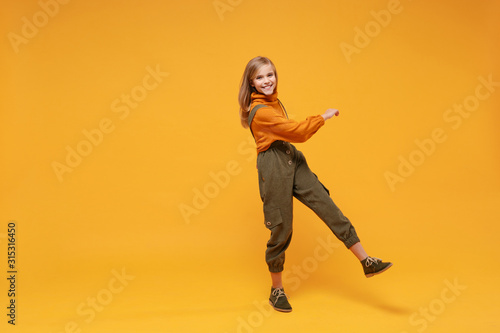Cheerful little blonde kid girl 12-13 years old in turtleneck, jumpsuit isolated on orange yellow background children studio portrait. Childhood lifestyle concept. Mock up copy space. Looking camera.