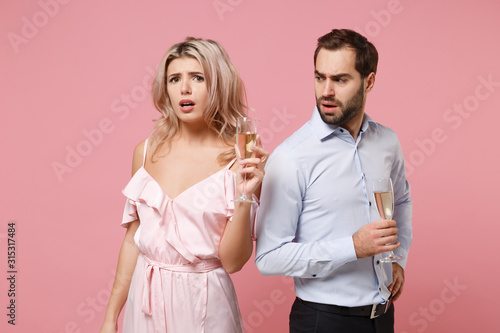 Puzzled young couple two guy girl in party outfit celebrating posing isolated on pastel pink background. People lifestyle Valentine's Day Women's Day birthday holiday concept. Hold glass of champagne.