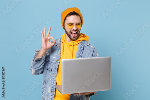 Funny young hipster guy in fashion jeans denim clothes posing isolated on pastel blue wall background. People lifestyle concept. Mock up copy space. Working on laptop pc computer, showing OK gesture.