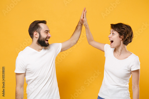 Laughing young couple friends bearded guy girl in white blank empty t-shirts isolated on yellow orange background. People lifestyle concept. Mock up copy space. Giving high five holding hands folded.