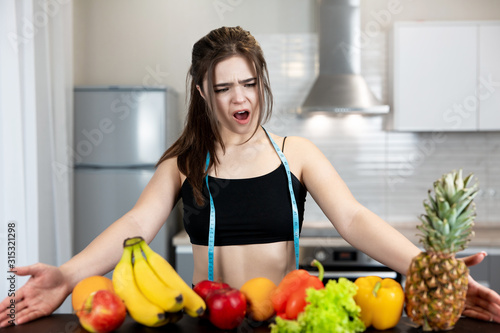 young fit woman with centimeter round neck wearing black sports top standing in the kitchen full of fruits looks shocked dietology and nutrition