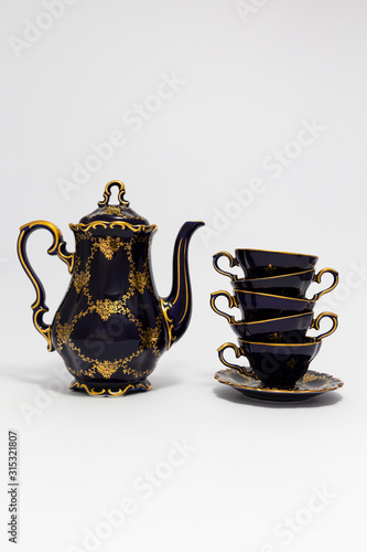 Closeup of a beautiful cobalt blue colored vintage porcelain tea set with golden floral pattern on white background. The set includes a tea pot, and a stack of tea cups.