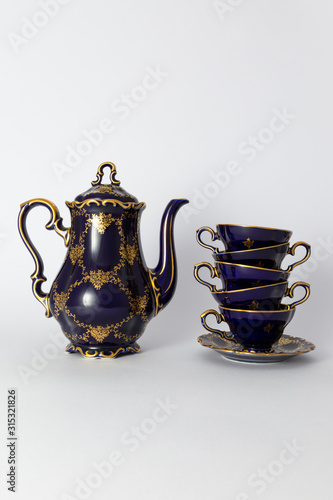 Closeup of a beautiful cobalt blue colored vintage porcelain tea set with golden floral pattern on white background. The set includes a tea pot, and a stack of tea cups.