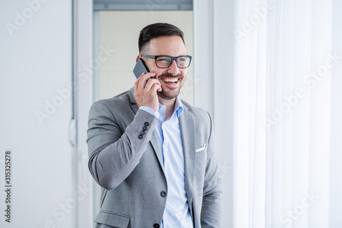 Attractive smiling unshaven caucasian businessman in suit and with eyeglasses using smart phone for business call while standing next to window in his office.