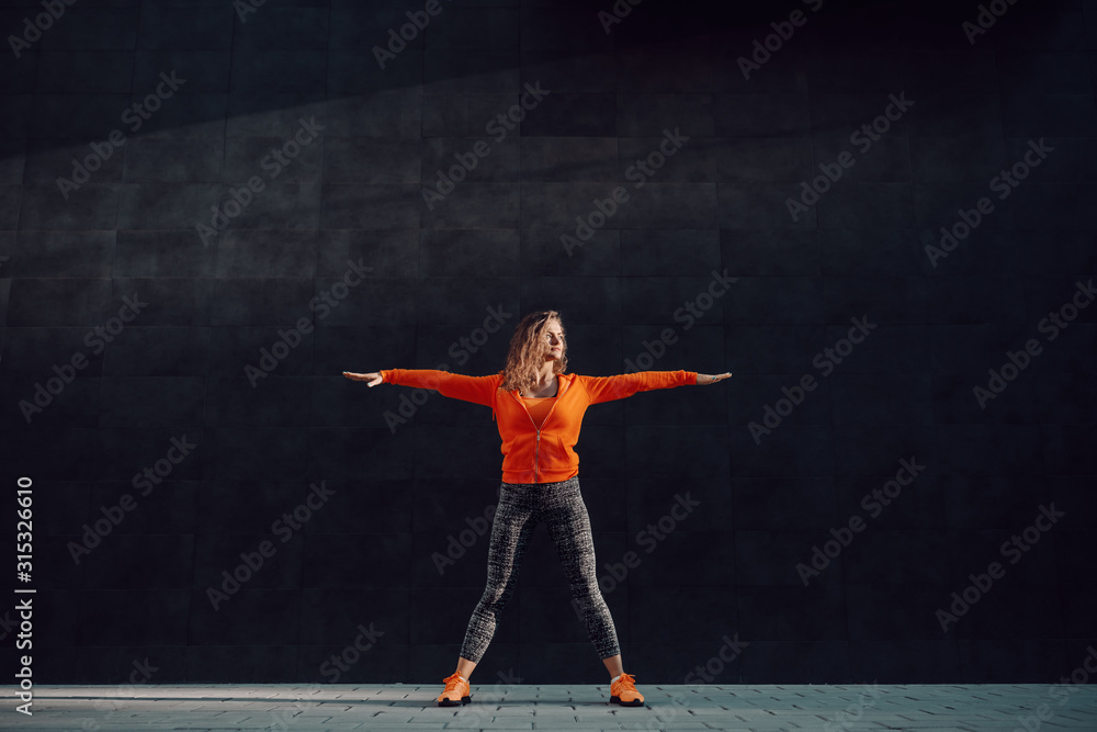 Full length of fit attractive caucasian woman in sportswear and with curly hair standing wide-legged and doing warm up exercises in front of black wall.