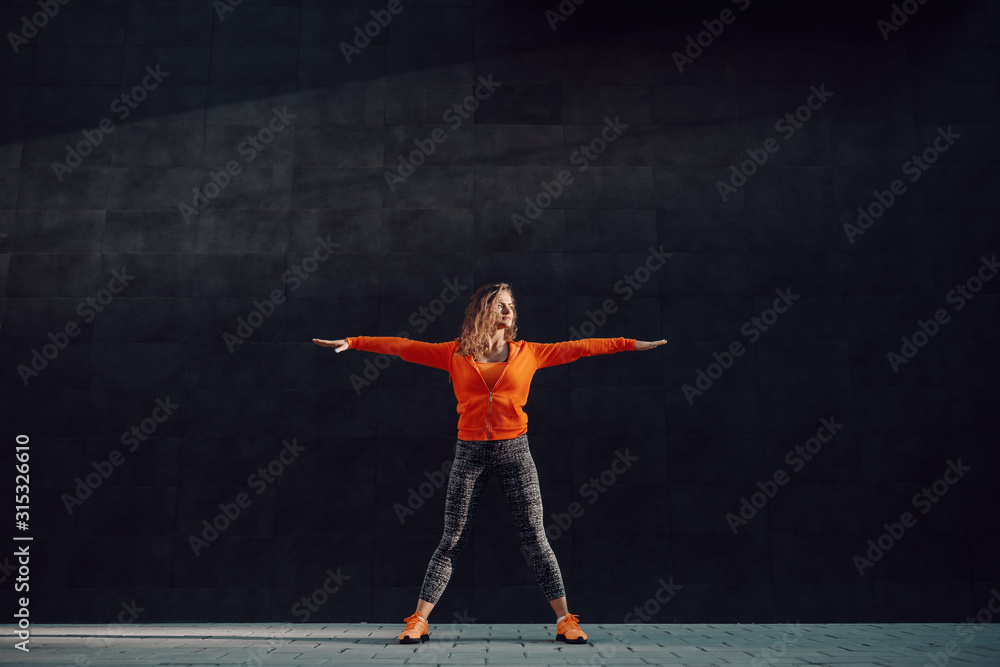 Full length of fit attractive caucasian woman in sportswear and with curly hair standing wide-legged and doing warm up exercises in front of black wall. Full length of fit attractive caucasian woman in sportswear and with curly hair standing wide-legged and doing warm up exercises in front of black wall.
