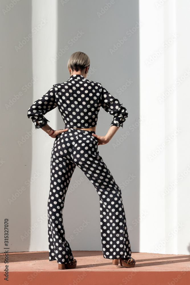 Fototapeta premium beautiful caucasian girl in a black pantsuit with white spots, pants and a shirt, high heel. Short haircut, bright makeup. Cool Posing at a white building with columns