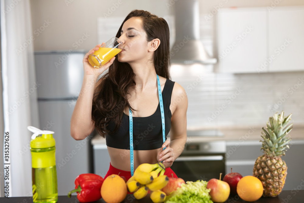 Fototapeta premium fit woman with centimeter round neck wearing black sports top standing in kitchen full of fruits drinks orange juice from the glass dietology and nutrition