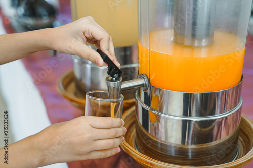 glass in girl's hand pouring cold orange juice from cooler, in soft focus