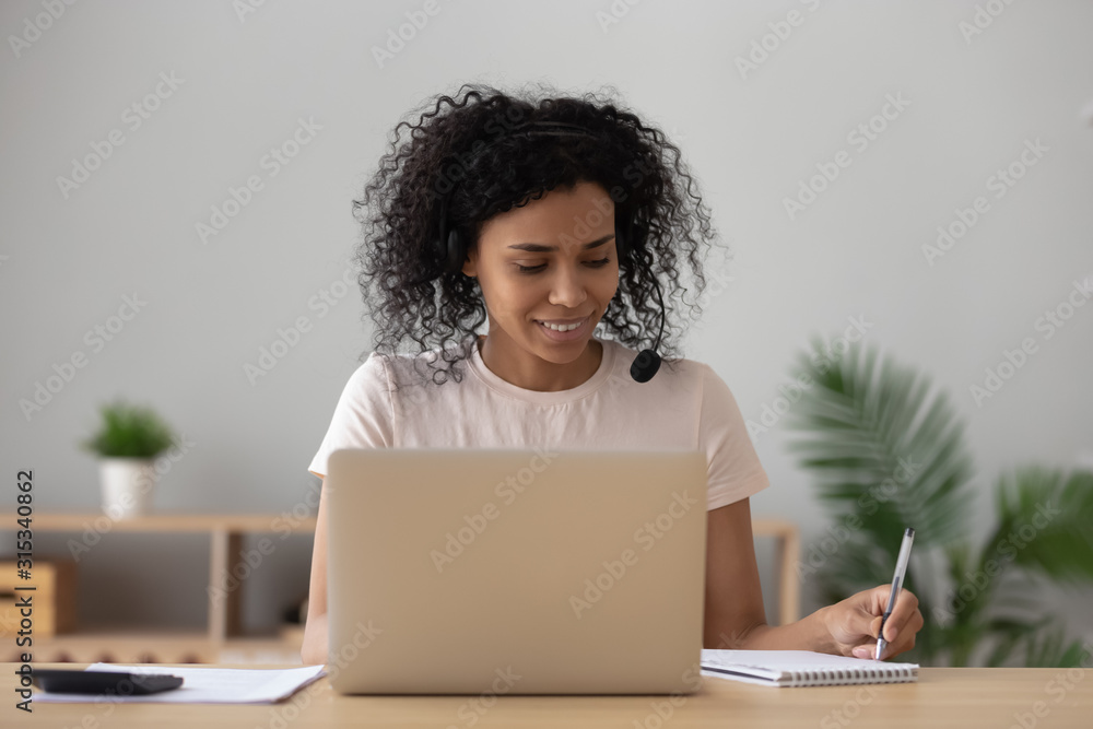African woman sitting at desk wearing headset hearing online webinar African woman sitting at desk wearing headset hearing online webinar