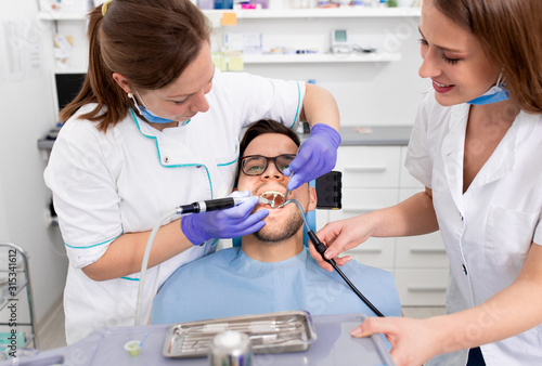 Female dentist with an assistant polishes teeth to a male patient in the dental clinic.