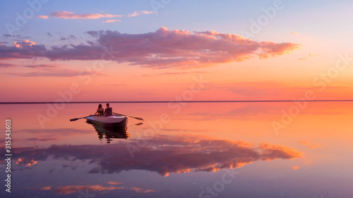 A couple in love look at beautiful sunset in a rowing boat on the lake. Pink sky and vanilla clouds. Romantic scene - lovers ride a boat in nature during sunset. Amazing landscape with people