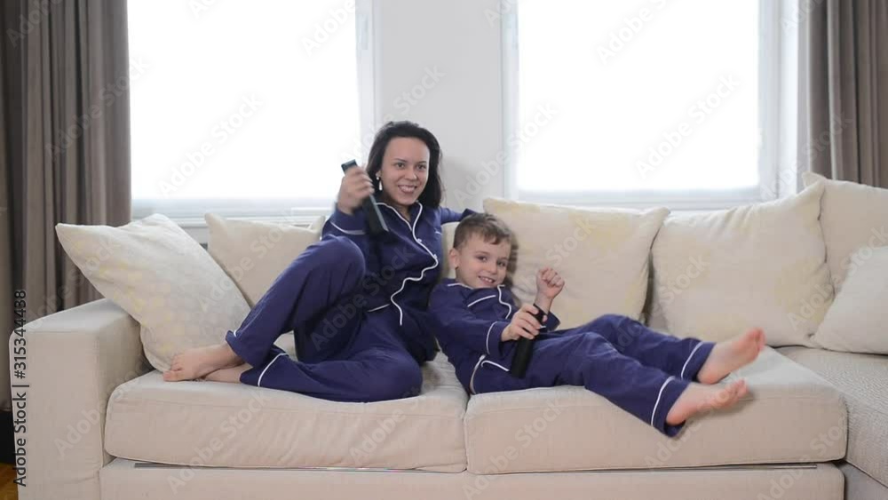 Happy young mother lying on bed and watching tv with her son. Both a wearing the same blue pyjamas, happy time on bed Happy young mother lying on bed and watching tv with her son. Both a wearing the same blue pyjamas, happy time on bed