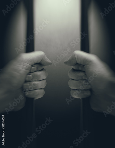 In jail concept with male hands holding jail bars, light in background room, first person view, toned image