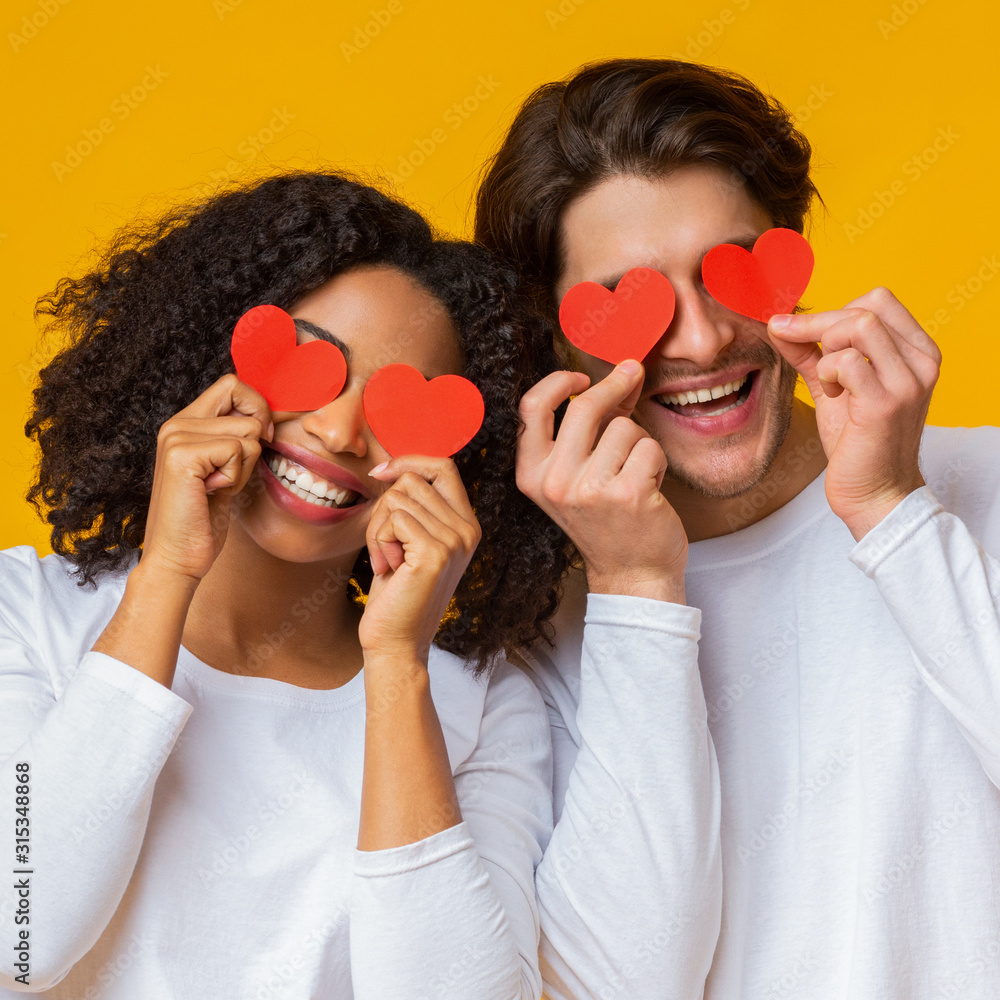 Romantic multiracial couple holding red paper hearts over eyes and smiling