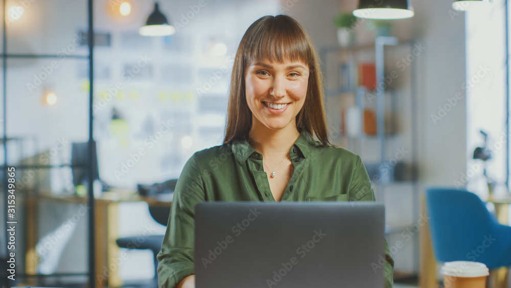 Young Beautiful Brunette Works on a Laptop Computer in Cool Creative Agency in a Bright Loft Office. She Smiles and Laughs on Camera. Young Beautiful Brunette Works on a Laptop Computer in Cool Creative Agency in a Bright Loft Office. She Smiles and Laughs on Camera.