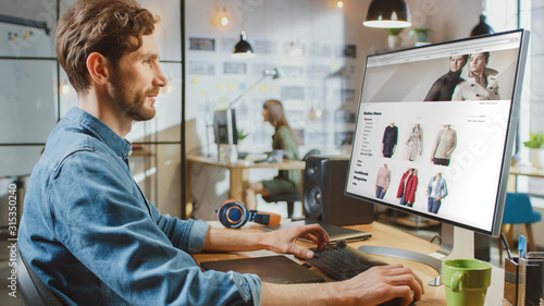 Beautiful Male Scrolls Through Online Fashion Store on His Personal Computer with Big Display. He Works in a Cool Office Loft. Other Female Creative Colleague Works in the Background. photo