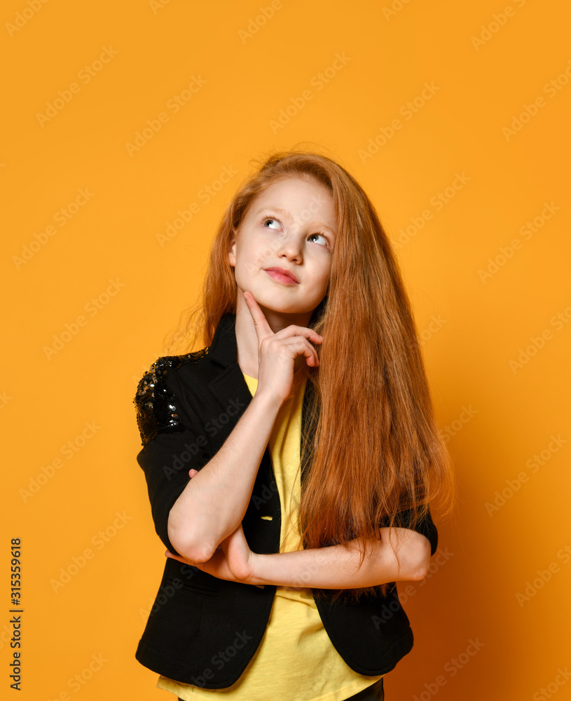 Ginger teenage child in black jacket and pants, yellow t-shirt. She is looking thoughtful, posing on orange background. Close up