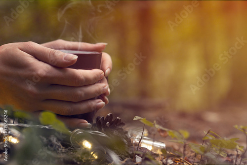 Young girl holding steaming cup of tea, surrounded by trees in the forest and warm autumn light. Cozy atmosphere, autumn vibes concept.