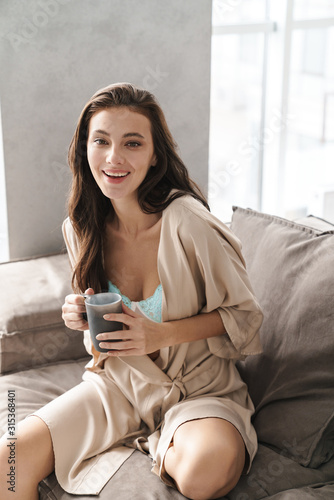 Image of young woman sitting on sofa at home and drinking tea in morning