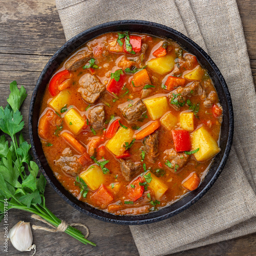 Goulash, beef stew or bogrash soup with meat, vegetables and spices in cast iron pan on wooden table. Hungarian cuisine. Rustic style. Top view. photo