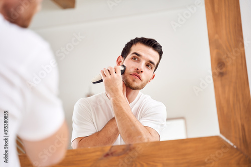 Young male in white T-shirt shaves while standing near mirror in bath