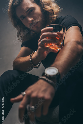 selective focus of fashionable businessman in black outfit with whiskey near grey wall