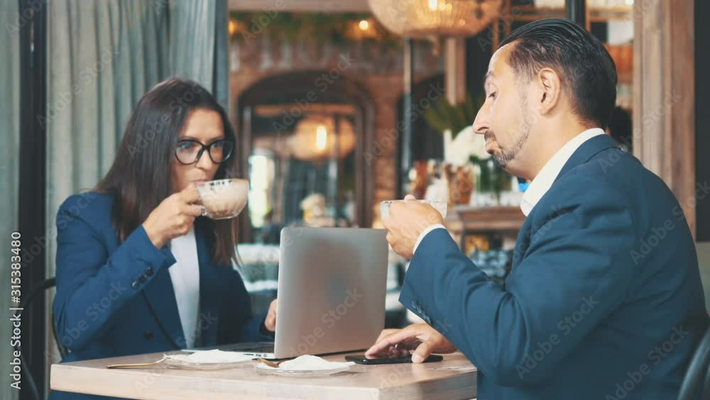 Two business people have business lunch. The man and woman in a restaurant or a coffee shop are drinking coffee, and woman is working on laptop. They showing thumbs up. Copy space. Two business people have business lunch. The man and woman in a restaurant or a coffee shop are drinking coffee, and woman is working on laptop. They showing thumbs up. Copy space.