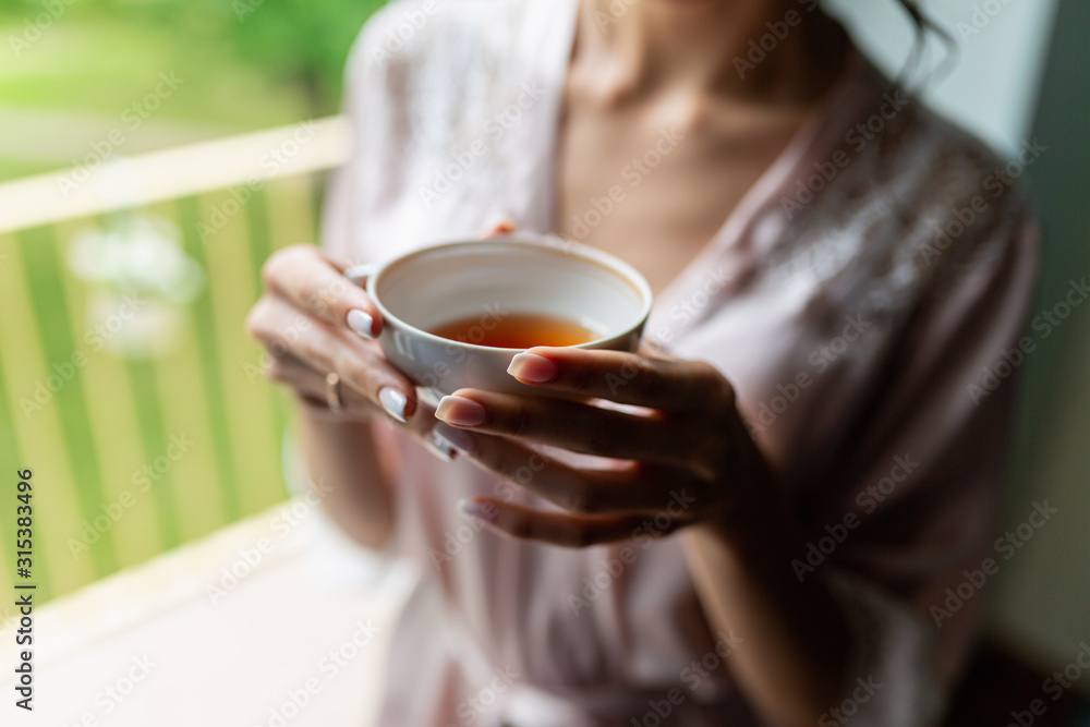 Close-up of a beautiful Cup of tea with a drawing in the hands of a girl in a soft pink robe. Cup in women's hands on the background of a bust in a white coat in the morning in a cozy hotel room Close-up of a beautiful Cup of tea with a drawing in the hands of a girl in a soft pink robe. Cup in women's hands on the background of a bust in a white coat in the morning in a cozy hotel room
