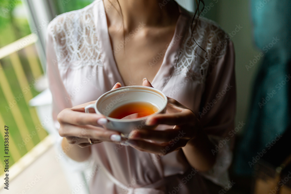 Close-up of a beautiful Cup of tea with a drawing in the hands of a girl in a soft pink robe. Cup in women's hands on the background of a bust in a white coat in the morning in a cozy hotel room Close-up of a beautiful Cup of tea with a drawing in the hands of a girl in a soft pink robe. Cup in women's hands on the background of a bust in a white coat in the morning in a cozy hotel room