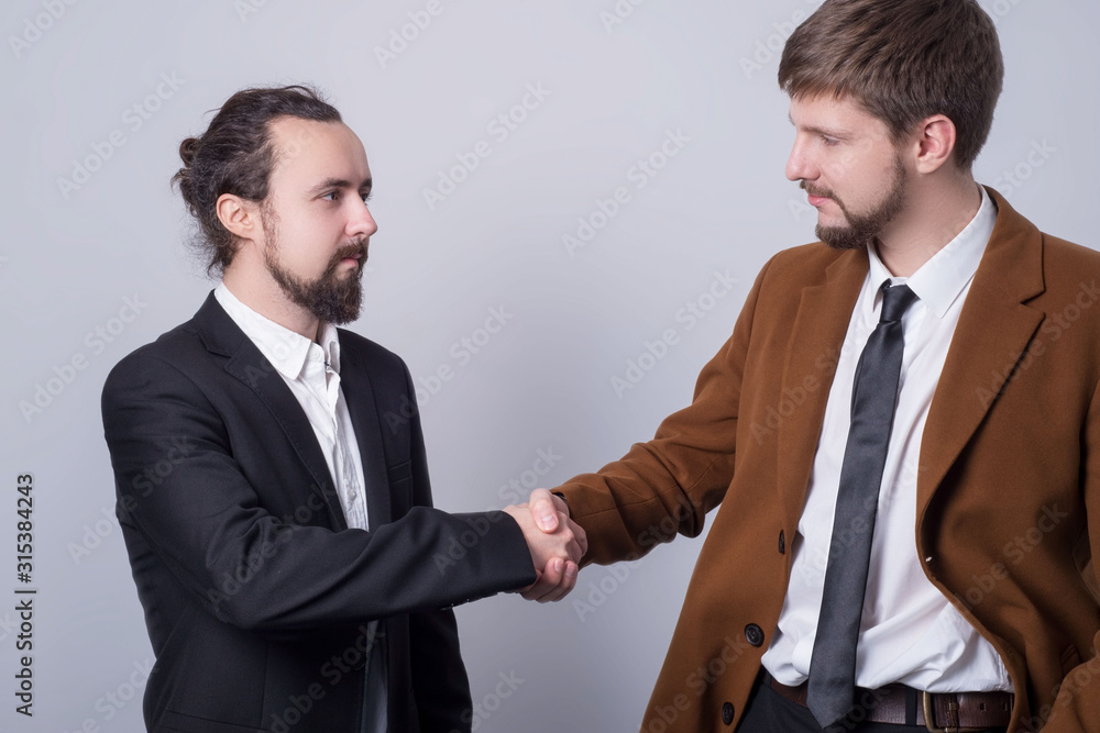 Portrait of two young people in business suits, smiling, shake hands, on a gray background. Simple business concept Portrait of two young people in business suits, smiling, shake hands, on a gray background. Simple business concept