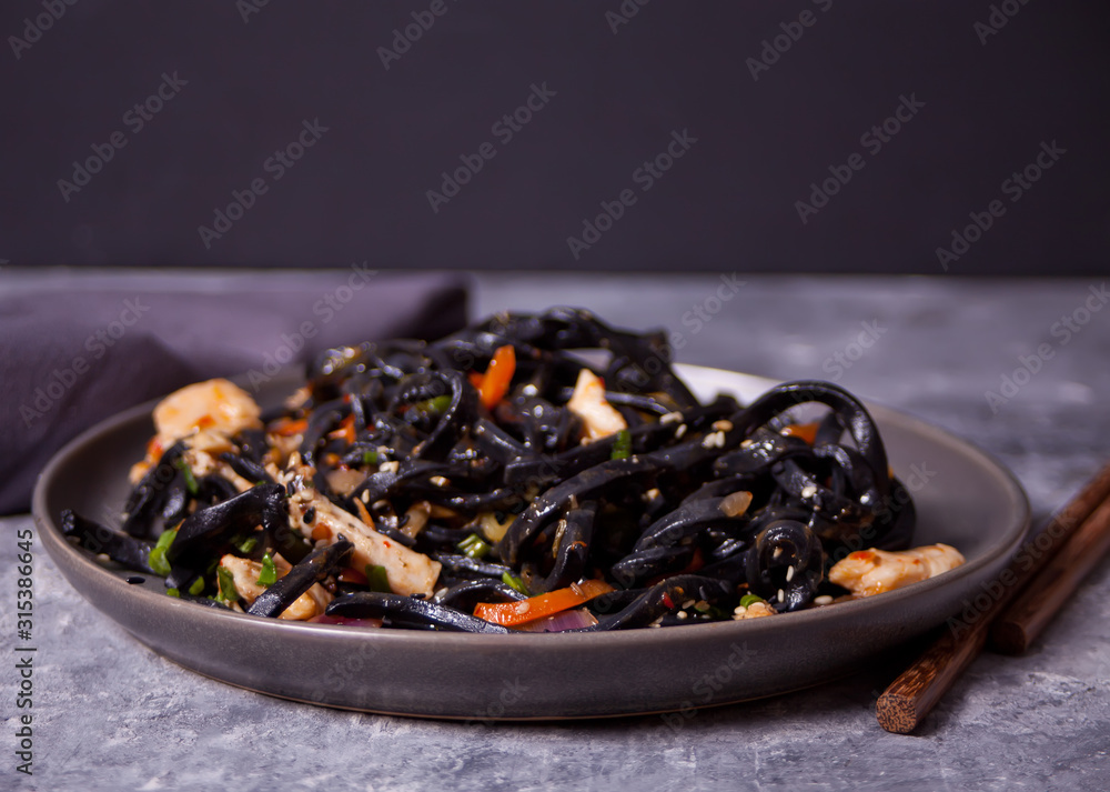 Black squid ink udon noodles with chicken meat on the black plate on dark stone background. Black squid ink udon noodles with chicken meat on the black plate on dark stone background.