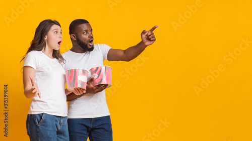 Surprised interracial friends holding popcorn buckets and pointing at copy space