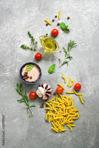 Raw pasta and ingredients for cooking. Fusilli, tomatoes, basil, olive oil, pink salt, pepper, rosemary and garlic on a gray concrete background. Top view, flat lay