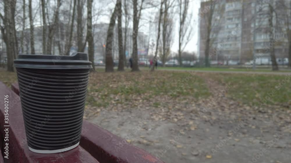 Close-up of a plastic cup with coffee standing on the street with a view of the park and apartment building.. People walking on a background. Slow motion. High resolution. Close-up of a plastic cup with coffee standing on the street with a view of the park and apartment building.. People walking on a background. Slow motion. High resolution.