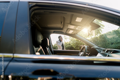 Photo of the car window on sunny day and successful African businessman walking while taking at the phone on the background. African american businessman in a suit speaking on smartphone.