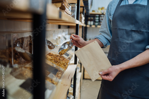 Mixed race male worker shop assistant filling paper bag with oat granola in bulk products in dispensers and food available at zero waste shop.