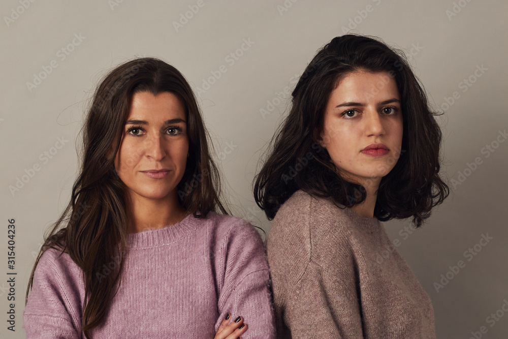 Satisfied young charming dark haired women wearing sweaters and looking at camera while standing together on gray background in studio Satisfied young charming dark haired women wearing sweaters and looking at camera while standing together on gray background in studio