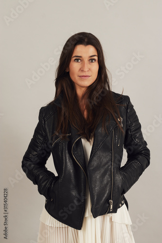 Thoughtful attractive dark haired woman in black biker jacket and looking at camera on gray background in studio photo