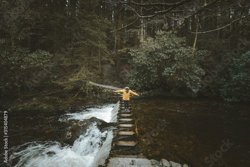 Side view of male tourist in bright orange jacket walking on footbridge and crossing river with water flowing through stepping stones in forest of Northern Ireland