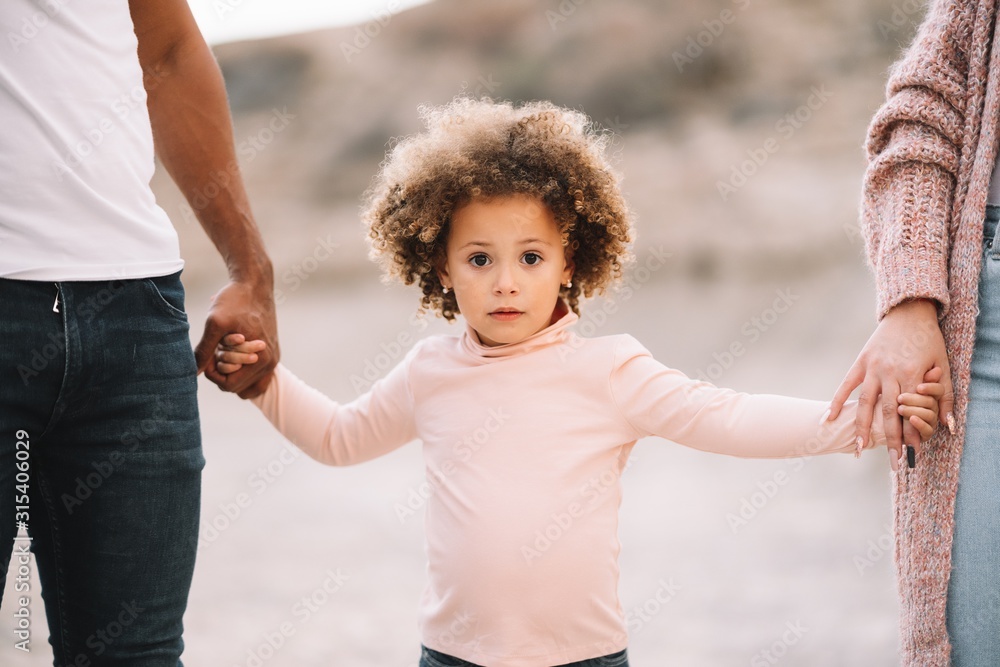 Cropped unrecognizable diverse mother father and curly child wearing bright casual clothes strolling on nature holding hands at daytime Cropped unrecognizable diverse mother father and curly child wearing bright casual clothes strolling on nature holding hands at daytime