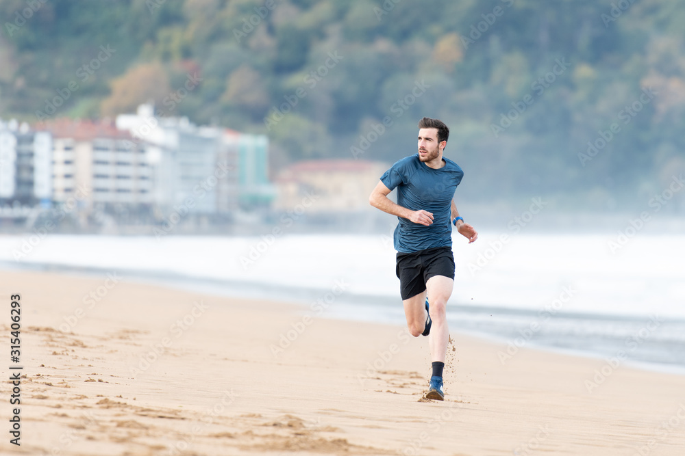 Bearded male athlete in active wear running during empty sandy beach with green mountains on blurred background Bearded male athlete in active wear running during empty sandy beach with green mountains on blurred background